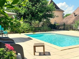 A swimming pool with a tiled deck, surrounded by greenery and a wooden fence.