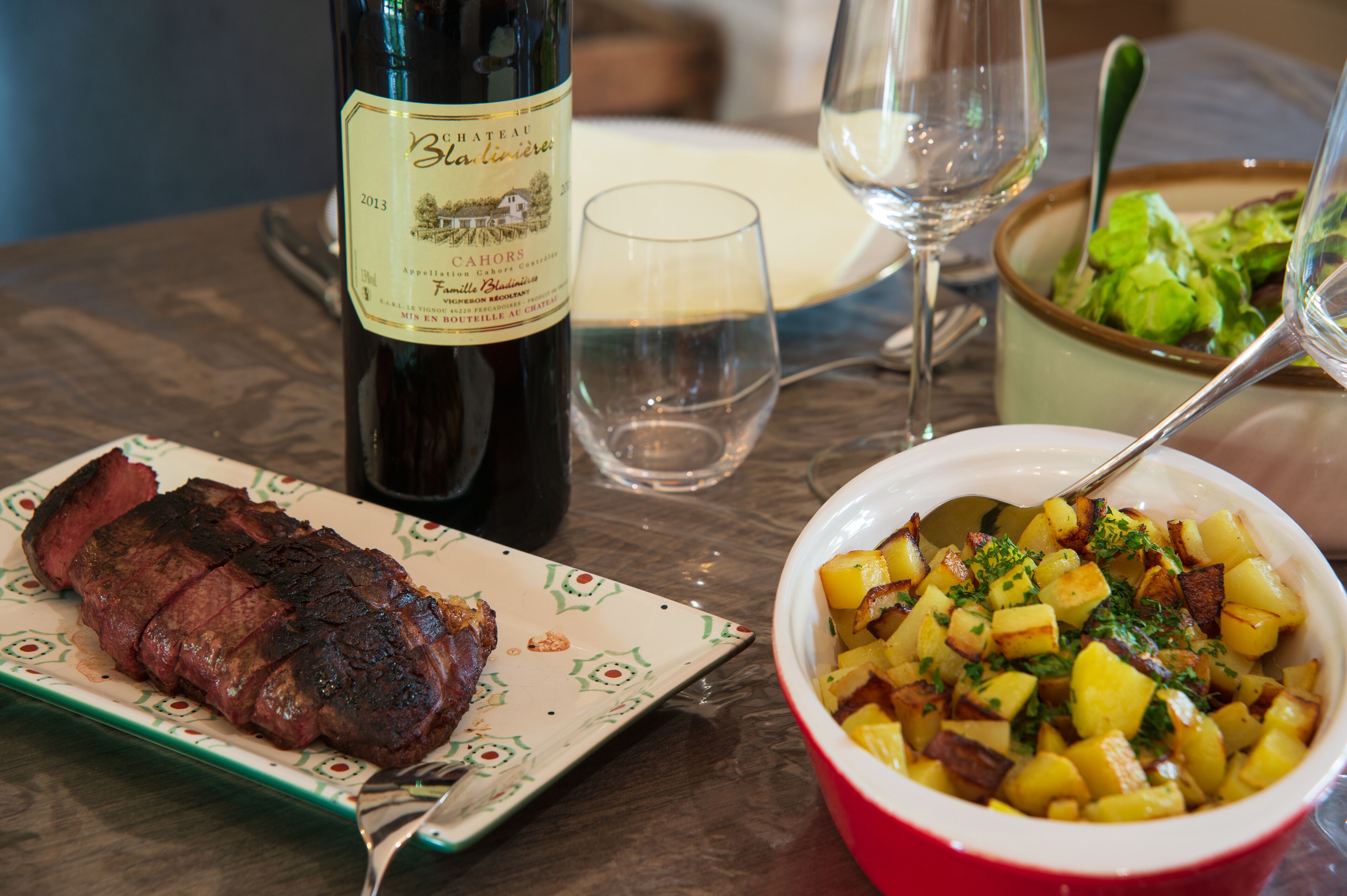 A dining table with a bottle of wine, a plate of steak, a bowl of potatoes, and a glass of water.