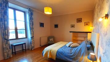 A bedroom with a bed, a bedside table, a chair, a window with curtains, and wall-mounted lights.