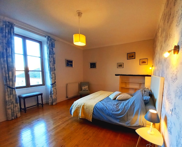 Un dormitorio con cama, una mesita de noche, una silla, una ventana con cortinas e iluminación en la pared.