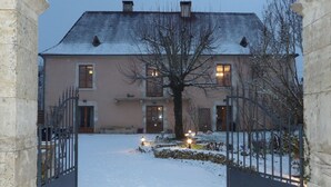 Entrada nevada a un edificio con un camino de acceso con portón, un árbol en primer plano y un edificio con varias ventanas y puertas.