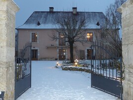 A snow-covered entrance to a building with a gated driveway, a tree in the foreground, and a building with multiple windows and doors.