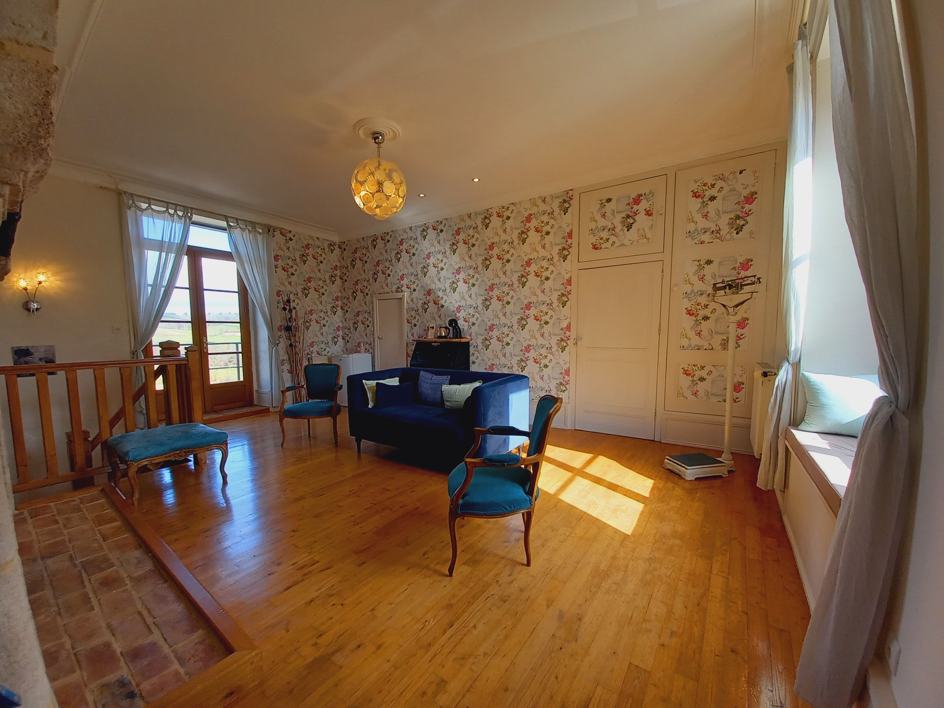 Un salón espacioso con un sofá azul, dos sillones, suelo de madera, papel tapiz floral y un gran ventanal con cortinas.
