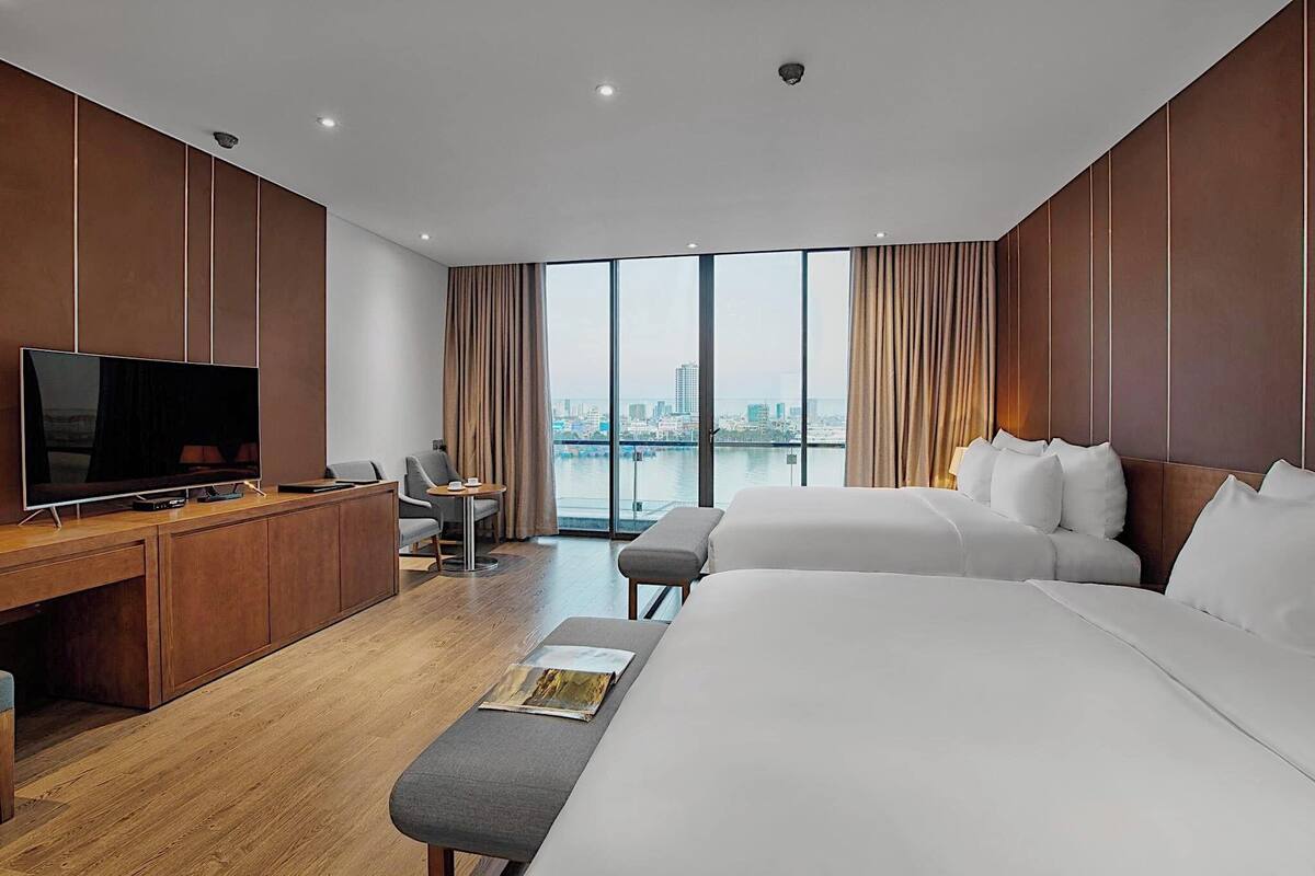 A hotel room with a large bed, a desk with a TV, a chair, and a view of the city through large windows.