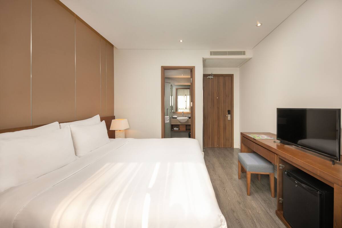 A hotel room with a large bed, a desk with a television, a small seating area, and a bathroom visible through an open door.