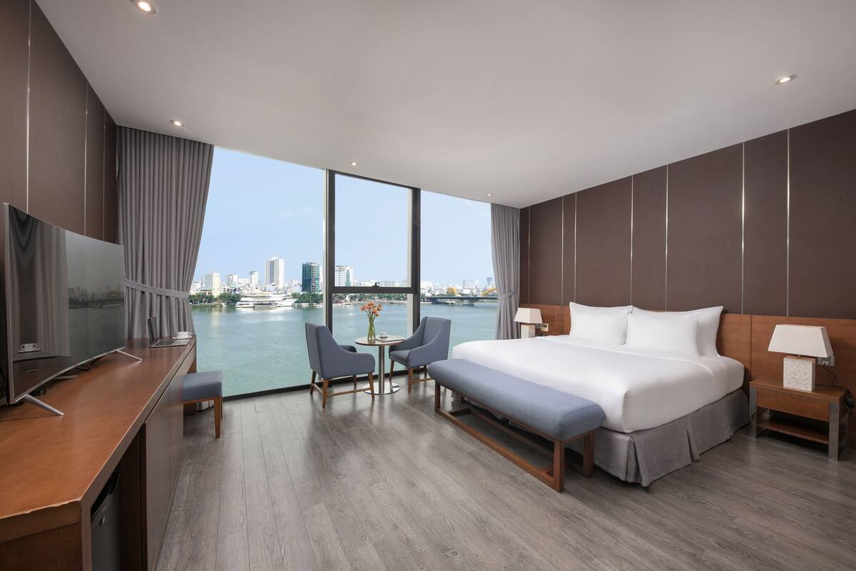 A modern hotel room with a large bed, a desk, a TV, and a view of a cityscape and water.