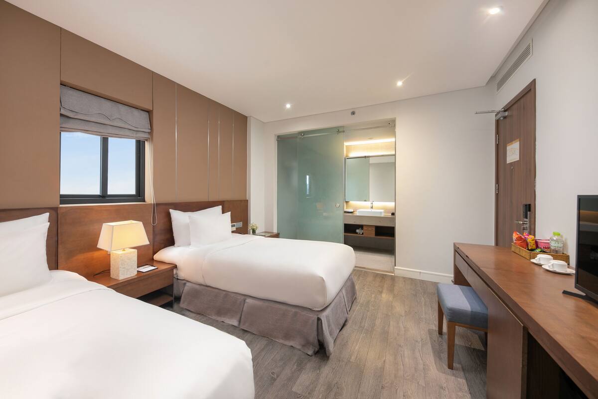 A hotel room with two beds, a desk, a television, and a bathroom with a glass door.