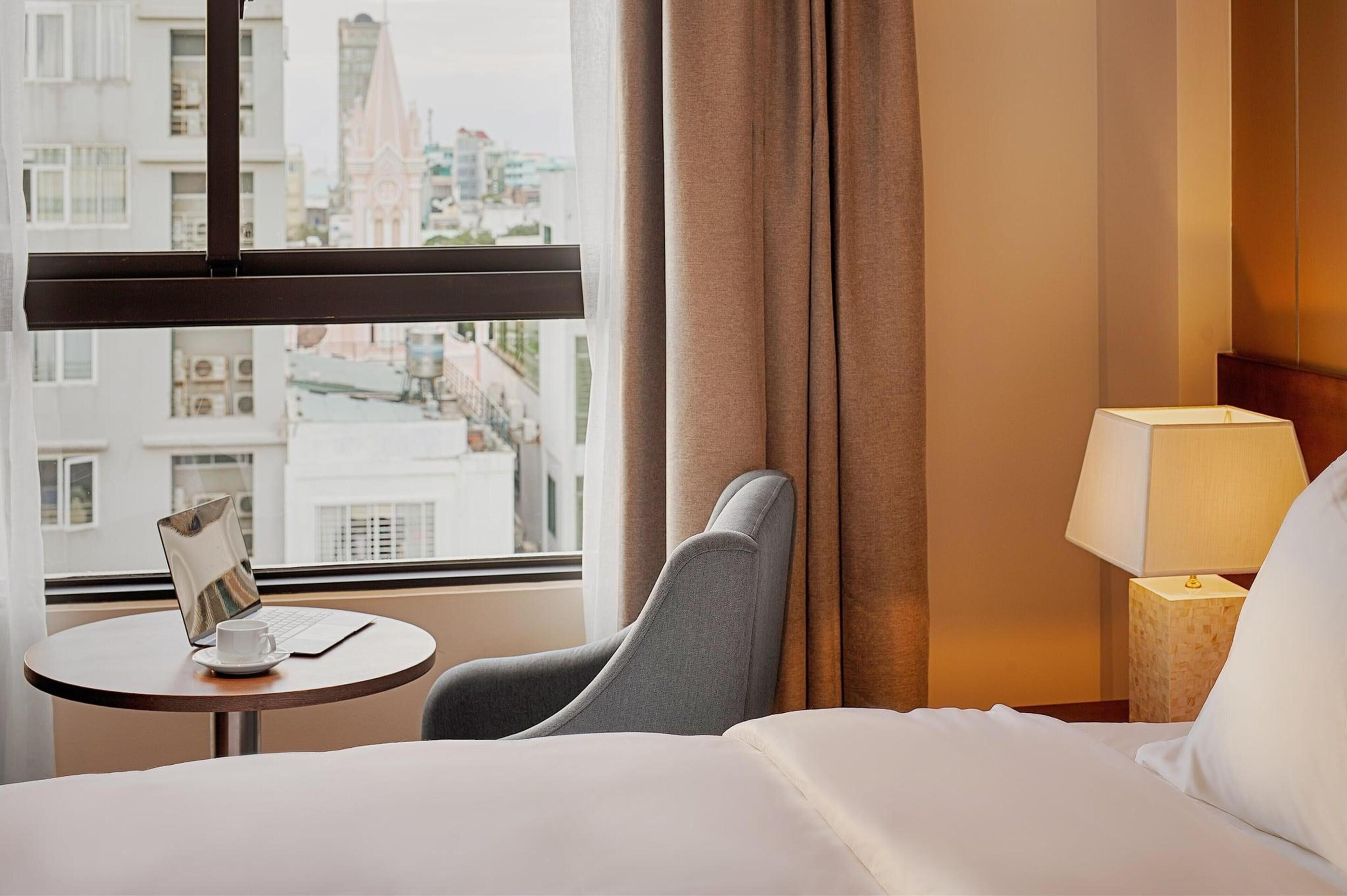 A hotel room with a bed, a desk with a laptop and a cup, a chair, a lamp, and a view of buildings through a window.