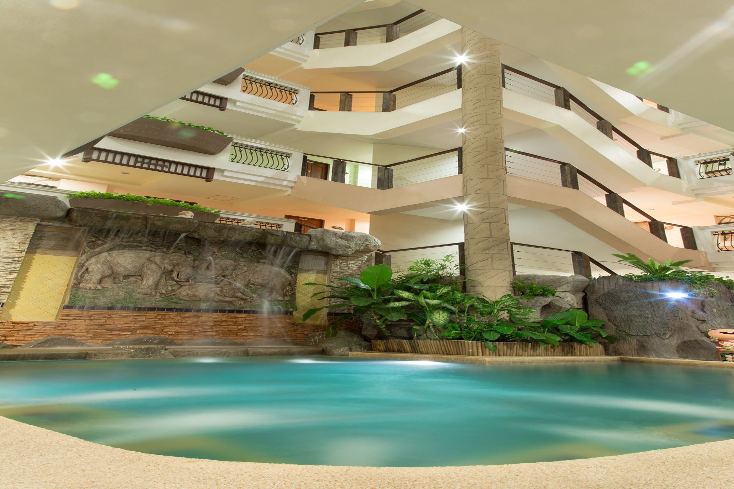 A multi-level indoor pool area with a waterfall feature, surrounded by plants and a stone wall.