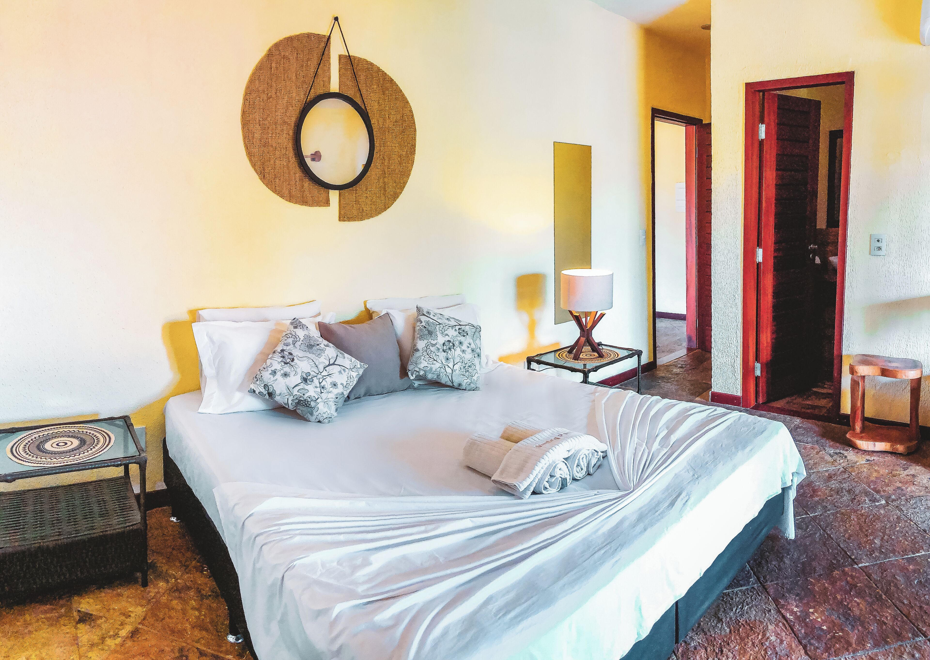 A neatly made bed with a white comforter and pillows, a small bedside table with a lamp, and a wall-mounted mirror.