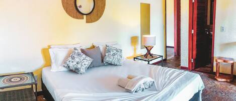A neatly made bed with a white comforter and pillows, a small bedside table with a lamp, and a wall-mounted mirror.