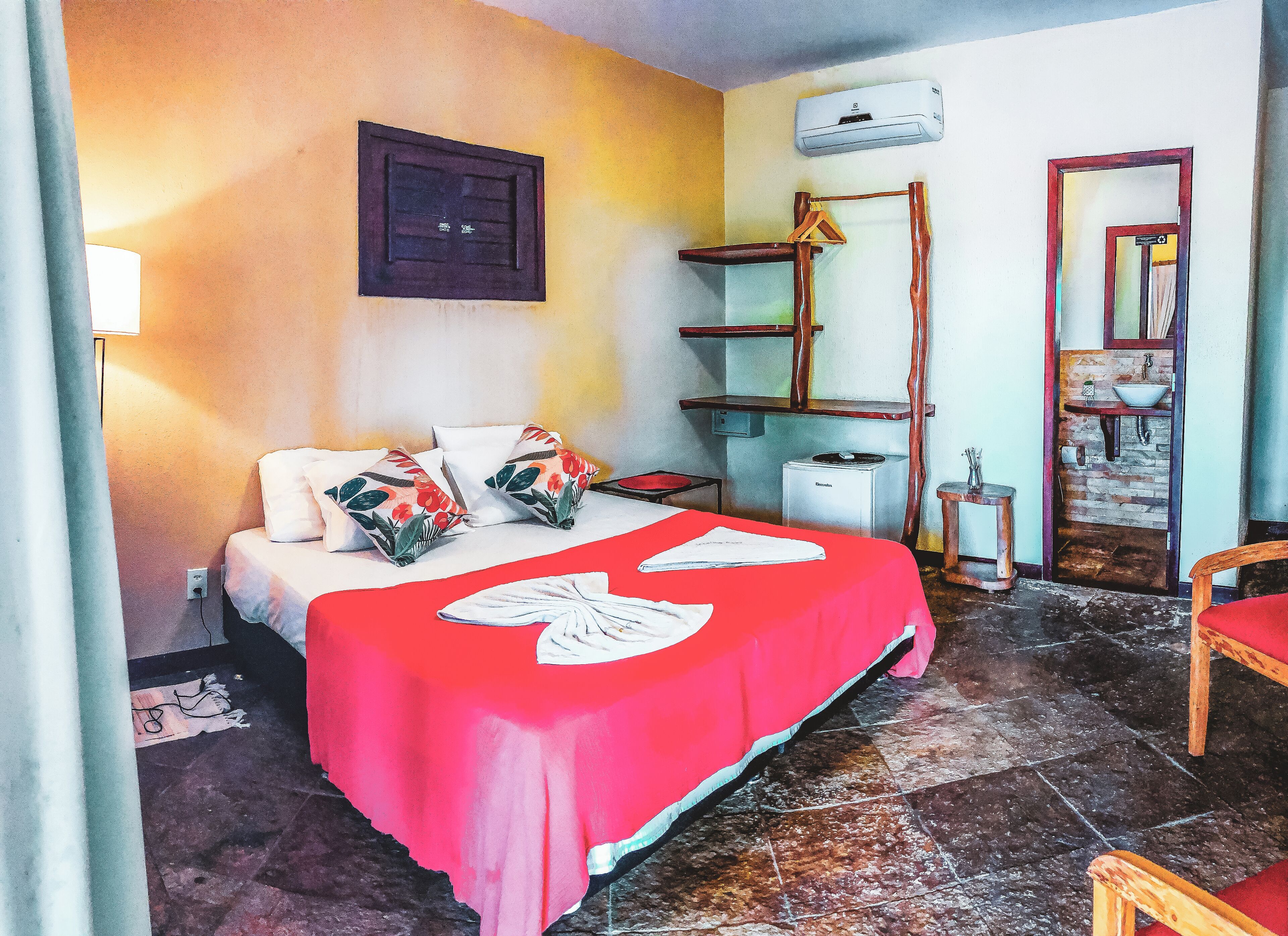 A hotel room with a bed, a red bedspread, a wall-mounted air conditioner, a wooden chair, and a bathroom visible through an open door.
