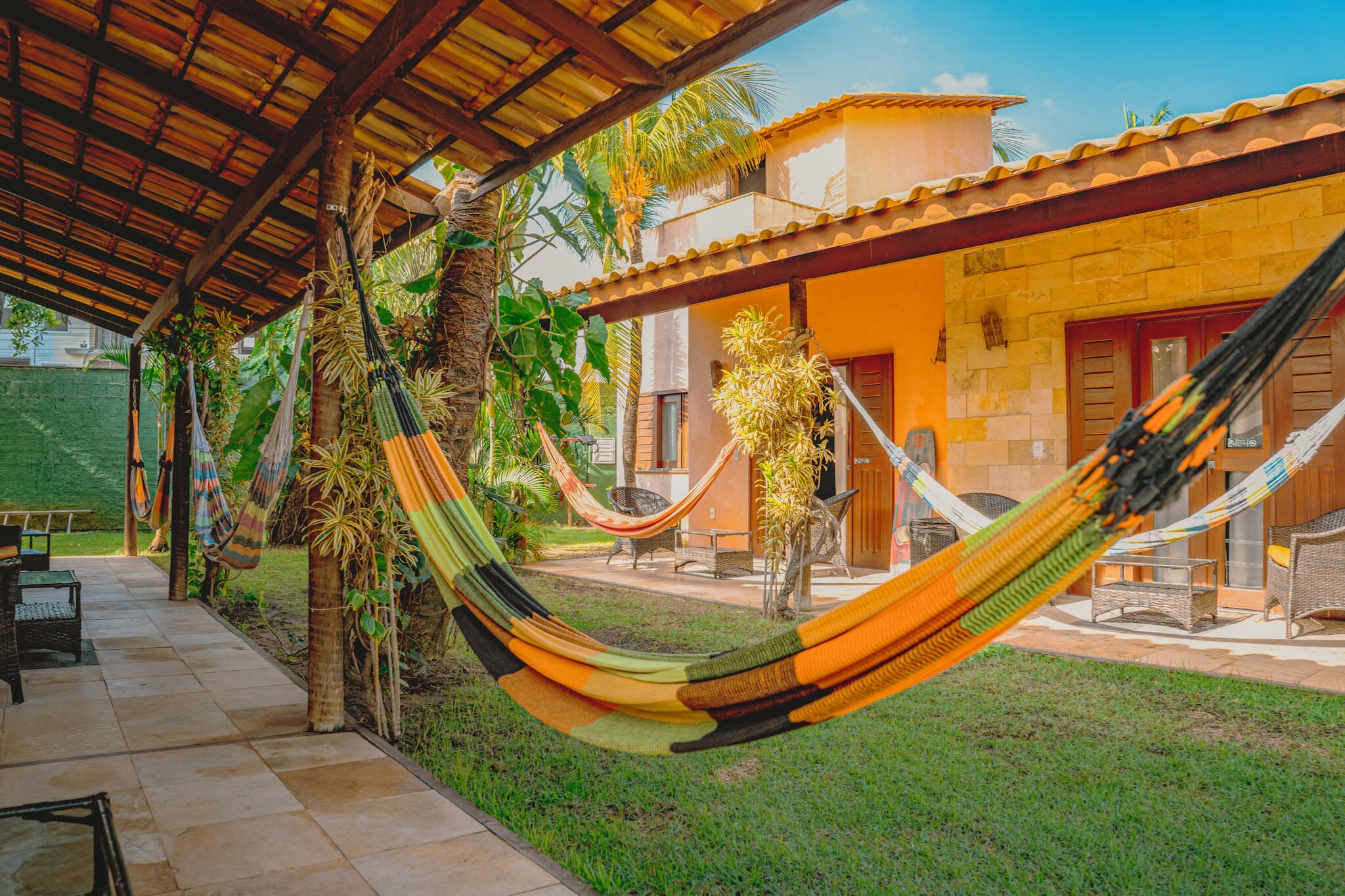 A hammock under a wooden pergola, a patio area with outdoor furniture, and a building with a tiled roof.