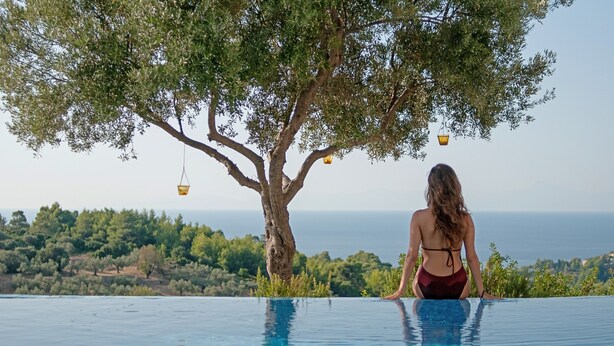Une femme est assise au bord d’une piscine à débordement, avec vue sur un arbre et l’océan.
