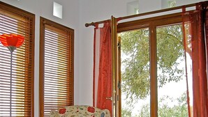 Une pièce comprenant un fauteuil à motifs floraux, une table ornée de vases rouges et offrant une vue sur la verdure à travers de grandes fenêtres équipées de stores en bois.