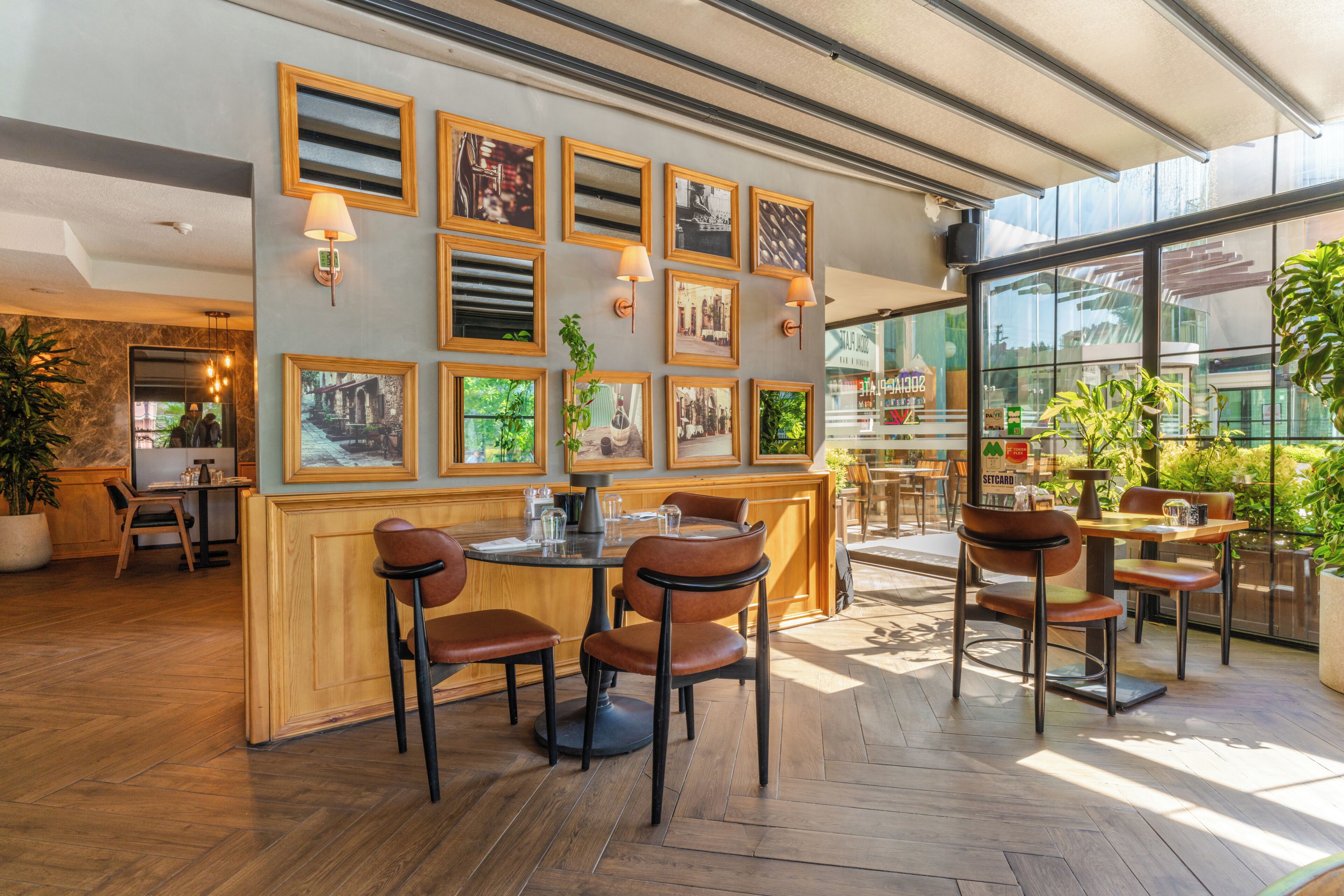 A restaurant interior with wooden furniture, framed pictures on the wall, and a large window offering a view.