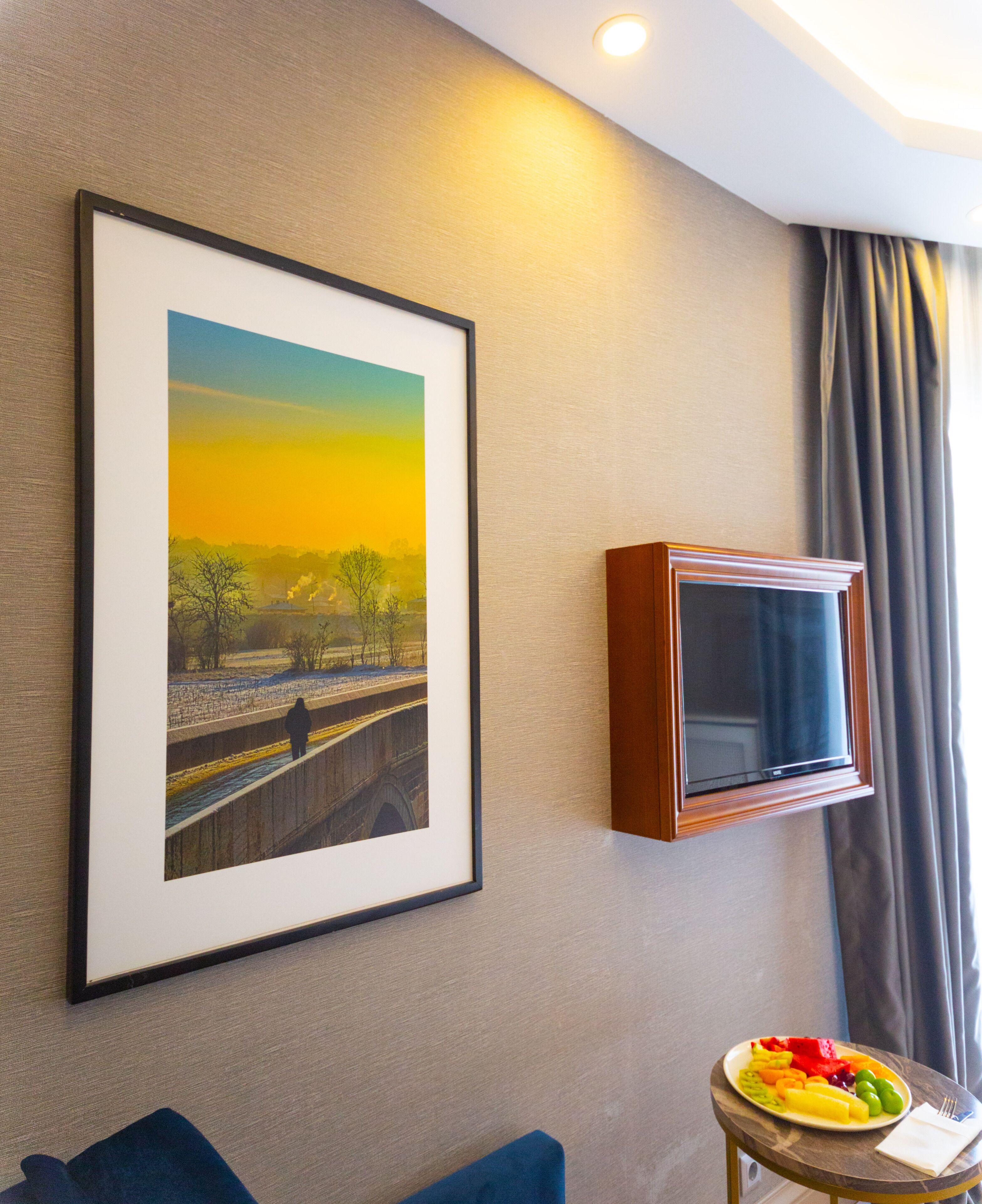 A framed landscape painting, a television, and a table with fruit and a napkin in a hotel room.