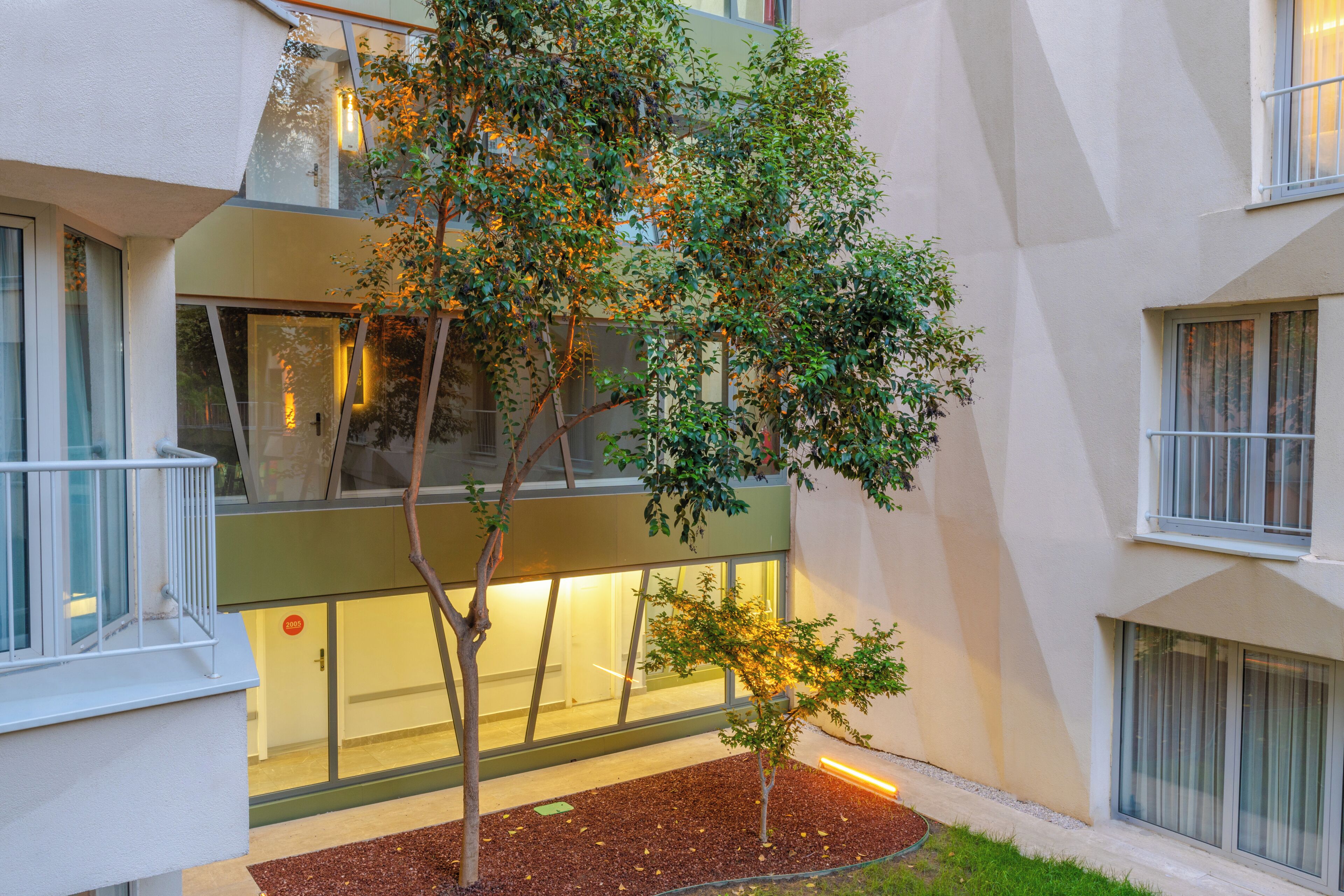 Un edificio moderno con fachada de vidrio, un pequeño árbol en el patio y una puerta visible con un cartel.