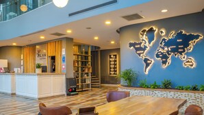 Un vestíbulo de hotel con un mostrador de recepción de madera, un mural de un mapa mundial, una estantería y un área de comedor con una mesa y sillas de madera.