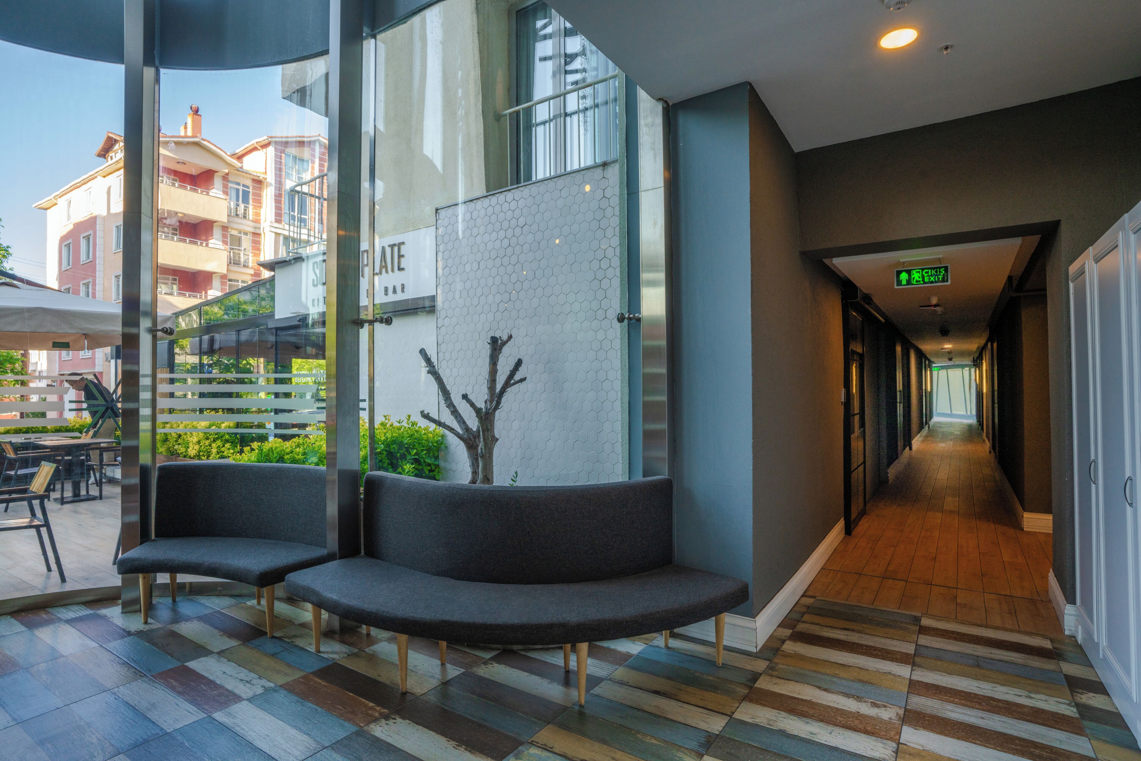 A modern hotel lobby with a checkered floor, a seating area with two sofas, a glass wall with a tree mural, and a hallway leading to an exit.