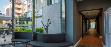 A modern hotel lobby with a checkered floor, a seating area with two sofas, a glass wall with a tree mural, and a hallway leading to an exit.