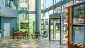 Un hall amplio con piso a cuadros, una zona de estar con un banco y sillas, una cafetería al fondo y una entrada con puerta de vidrio.