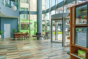 A spacious lobby with a checkered floor, a seating area with a bench and chairs, a coffee shop in the background, and a glass door entrance. - Alan Suites (Edirne)
