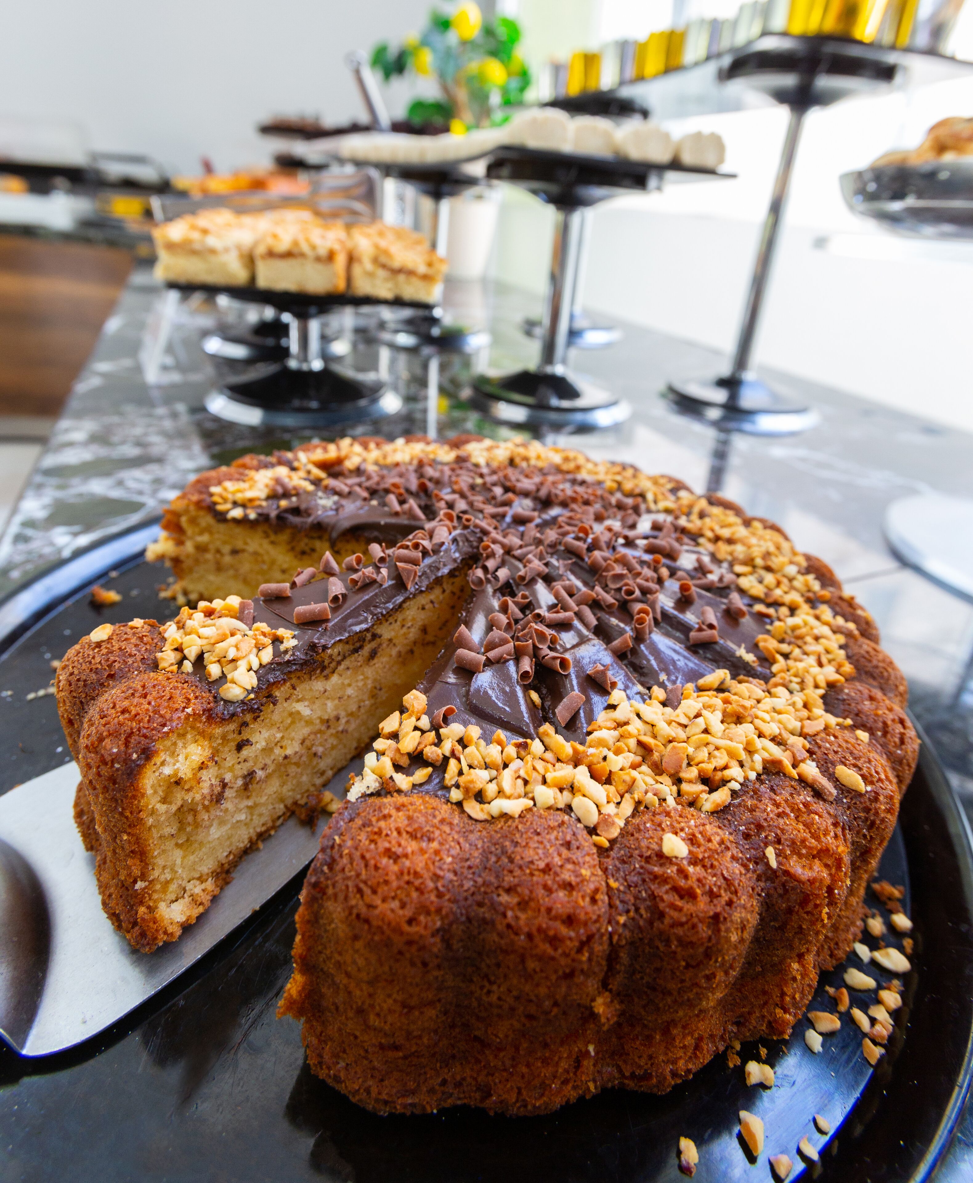 Un gâteau rond, une part ayant été coupée, garni de copeaux de chocolat et de noix, présenté sur une assiette noire.