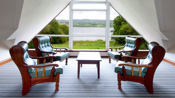 Ein Zimmer mit hohen Decken und einem großen Fenster mit Blick auf einen See und Hügel. Zwei Holzstühle mit grüner Polsterung und ein kleiner Tisch stehen in der Mitte.