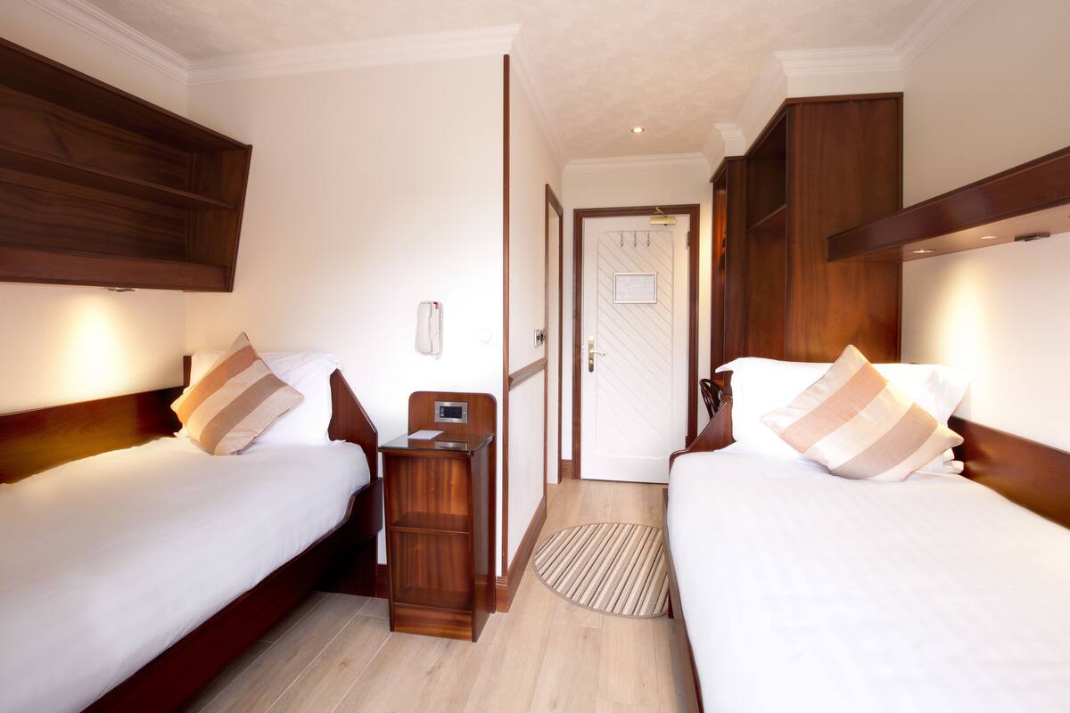 A hotel room with two beds, wooden headboards, a bedside table with a lamp, and a door leading to another room.