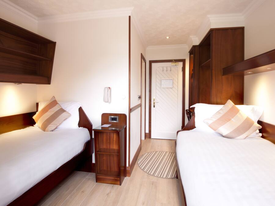Habitación de hotel con dos camas, cabeceros de madera, una mesita de noche con lámpara y una puerta que da a otra habitación.