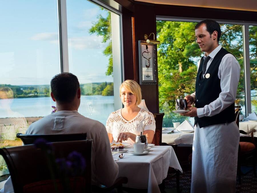 A restaurant with a view of a lake, a waiter serving tea, and a table set for two.