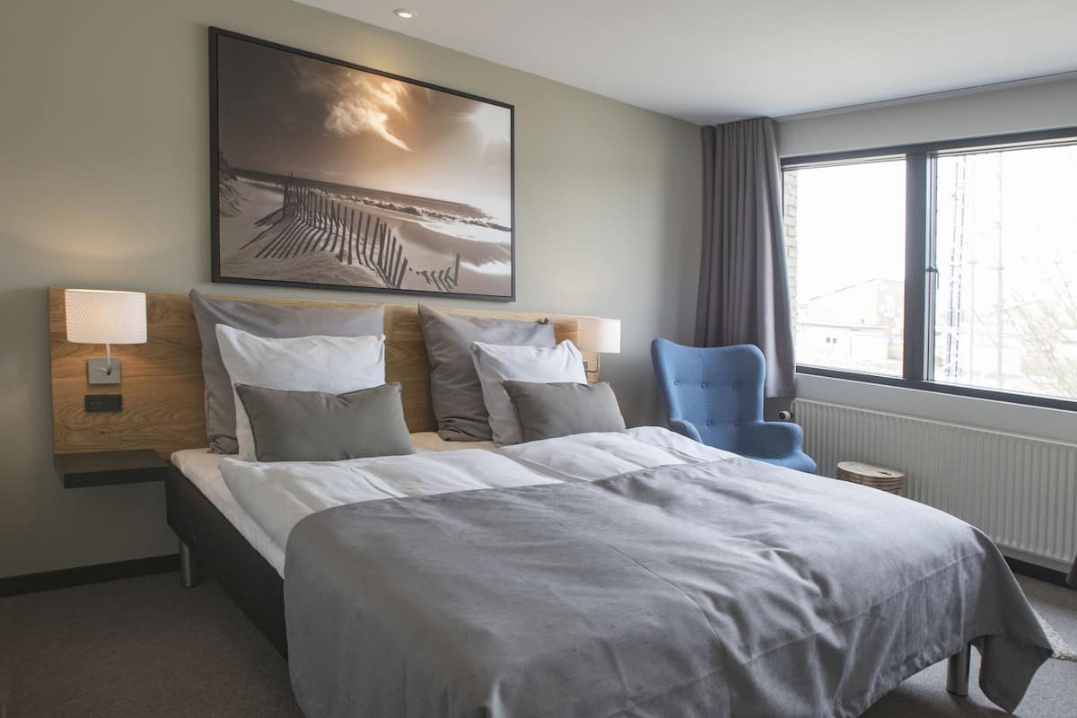 A hotel room with a large bed, two bedside tables with lamps, a chair, a large window, and a painting on the wall.