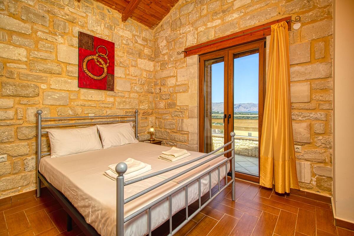 Un dormitorio con pared de piedra, cama con estructura metálica, techo de madera y vistas a través de un ventanal.