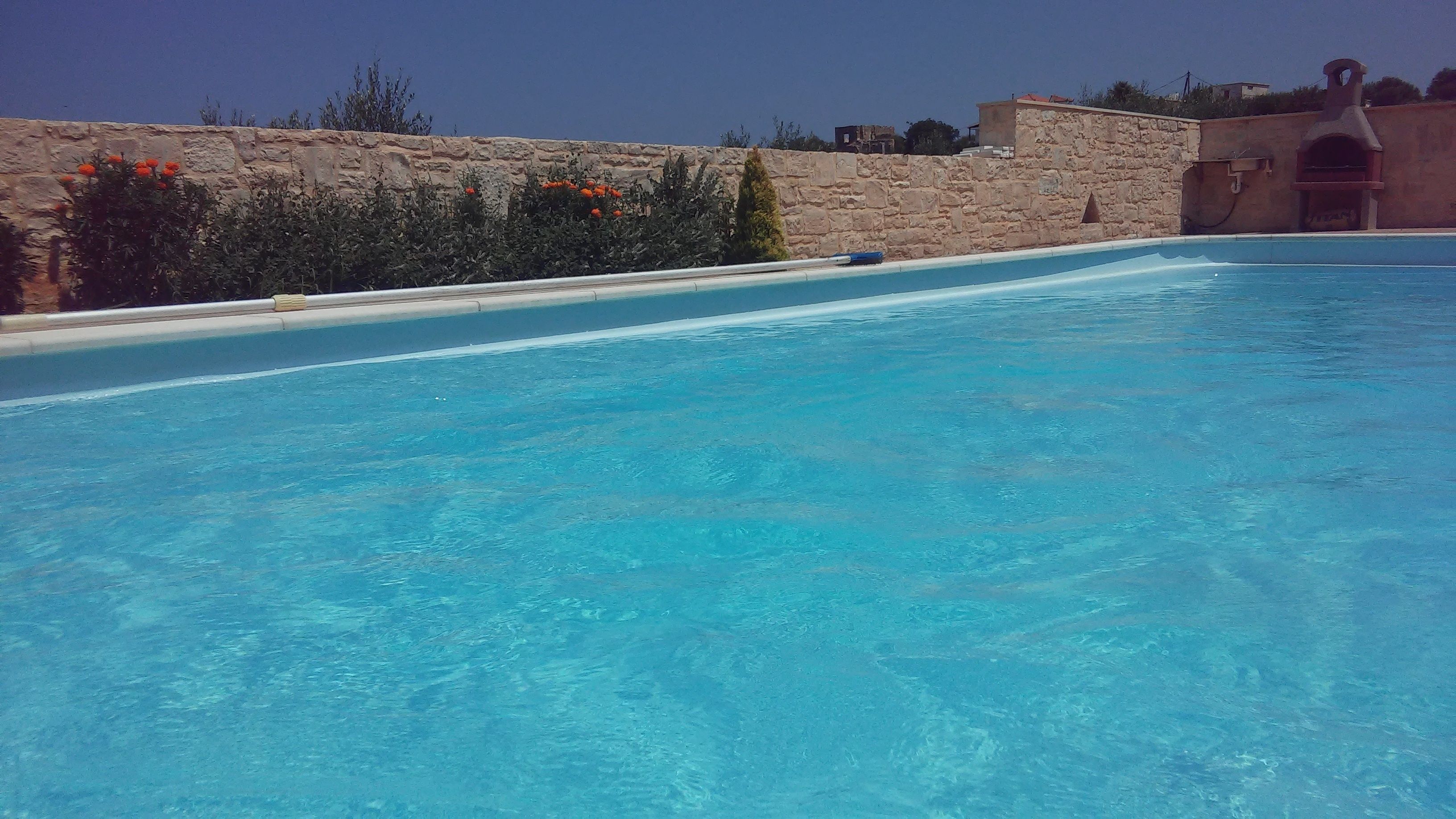 En klar, blå swimmingpool med stenvægge og en stenvæg med vegetation i baggrunden.