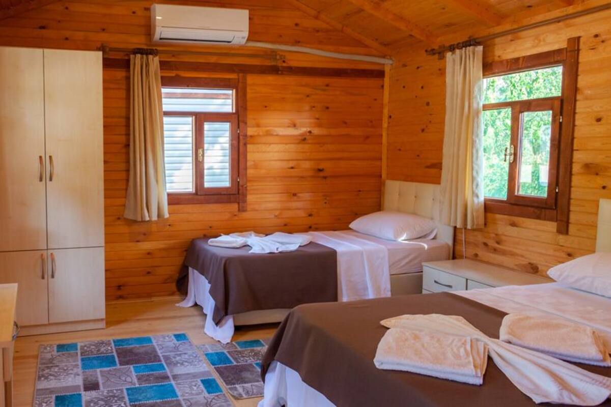 A wooden cabin room with two beds, a desk, a wardrobe, and a window with curtains.