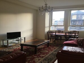 A living room with a television, a dining table with chairs, a chandelier, and a view of buildings through the window. - Menzies Apartment (Edinburgh)