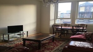 Un salon comprenant un téléviseur posé sur une table en verre, une table basse en bois, un tapis rouge à motifs, un canapé en cuir et un coin repas avec des chaises et une table.