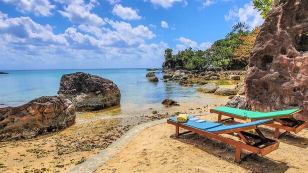 Una playa con dos sillas de madera para tomar el sol, un camino de piedra y una orilla rocosa.