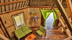 Una habitación con techo de paja, paredes de madera, un sofá, una silla, una pequeña mesa con una lámpara y un cuadro en la pared.