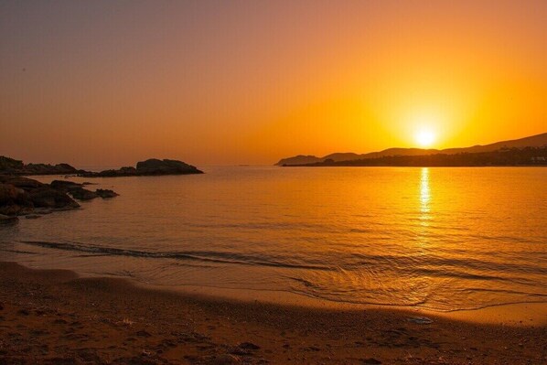 A sunset over a calm sea with a sandy beach and rocky outcrops.