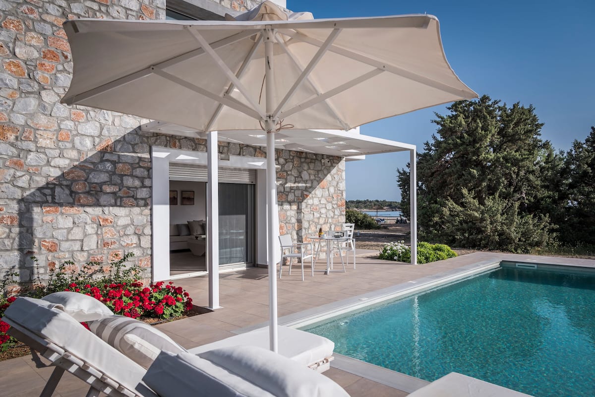 Zona junto a la piscina con pared de piedra, una silla de descanso, una mesa con sillas y una sombrilla.