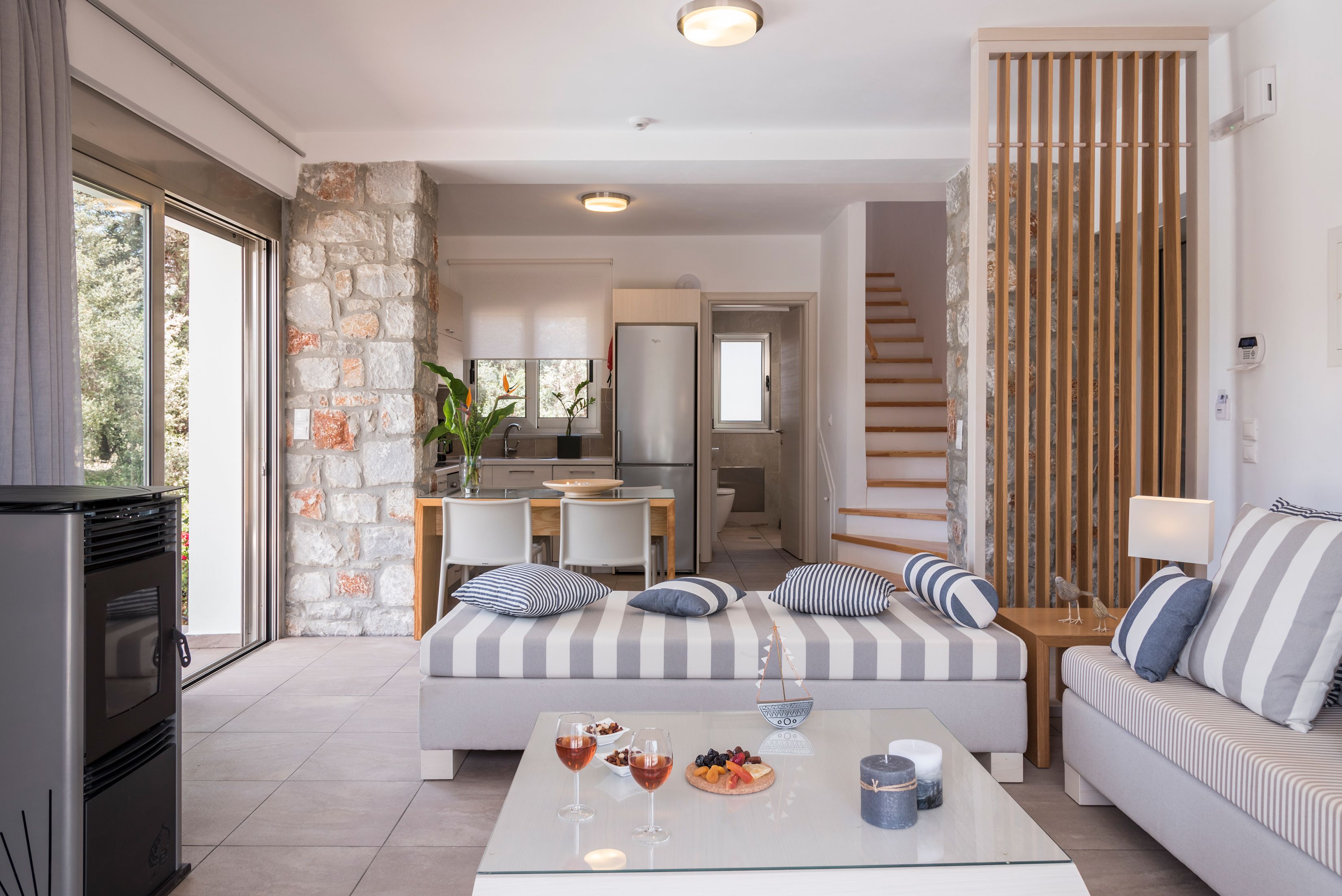 Un salon moderne avec un mur d’accent en pierre, un canapé avec des coussins à rayures, une table basse en verre sur laquelle se trouvent des plats et des boissons, et une cuisine en arrière-plan.
