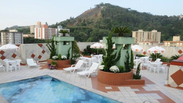 A rooftop pool area with white lounge chairs and umbrellas, surrounded by buildings and a hilly landscape. - Serra Negra Paladium Hotel (Serra Negra)