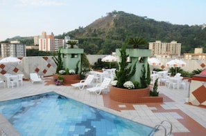 Un espace piscine aménagé sur le toit, avec des chaises longues blanches et des parasols, entouré de bâtiments et surplombant un paysage vallonné.