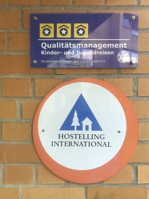 Zwei Schilder an einer Backsteinmauer. Das obere Schild ist blau mit gelben Sternen und Text auf Deutsch, das eine Organisation für Qualitätsmanagement im Bereich Kinder- und Jugendreisen kennzeichnet. Das untere Schild ist weiß mit einem blauen Dreieck und Text, das Hostelling International repräsentiert.