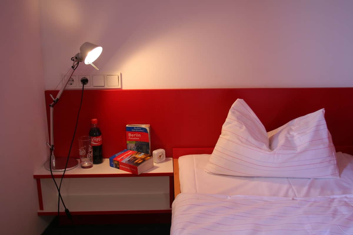A hotel room with a red headboard, a bedside lamp, a bottle of Coca-Cola, a book titled 'Berlin Potsdam', and a white pillow on the bed.