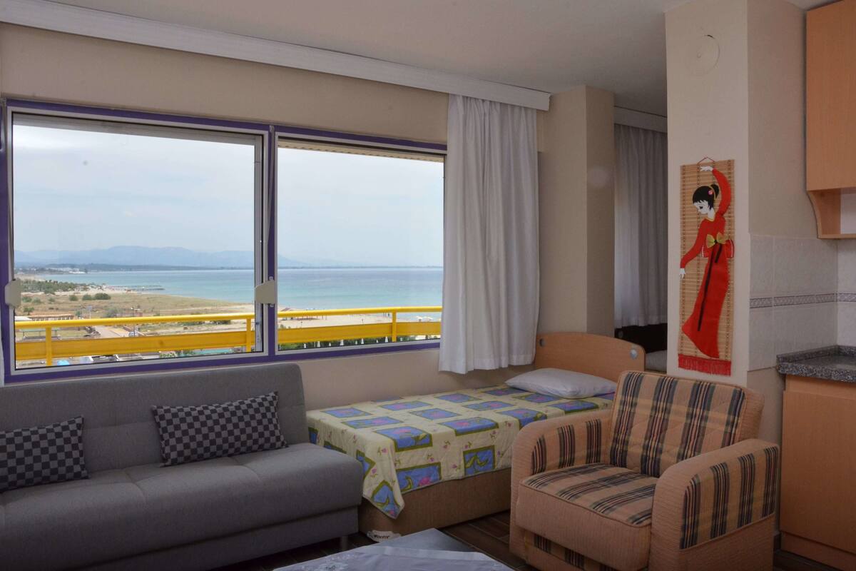 A hotel room with a bed, a sofa, a chair, a window with a view of the sea, and a small kitchen area.
