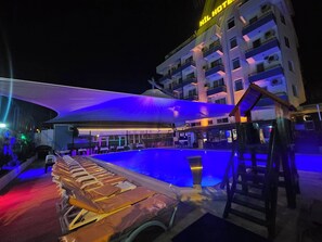 A hotel pool area with a covered seating area and a building in the background. - Nil Hotel (Ayvalik)