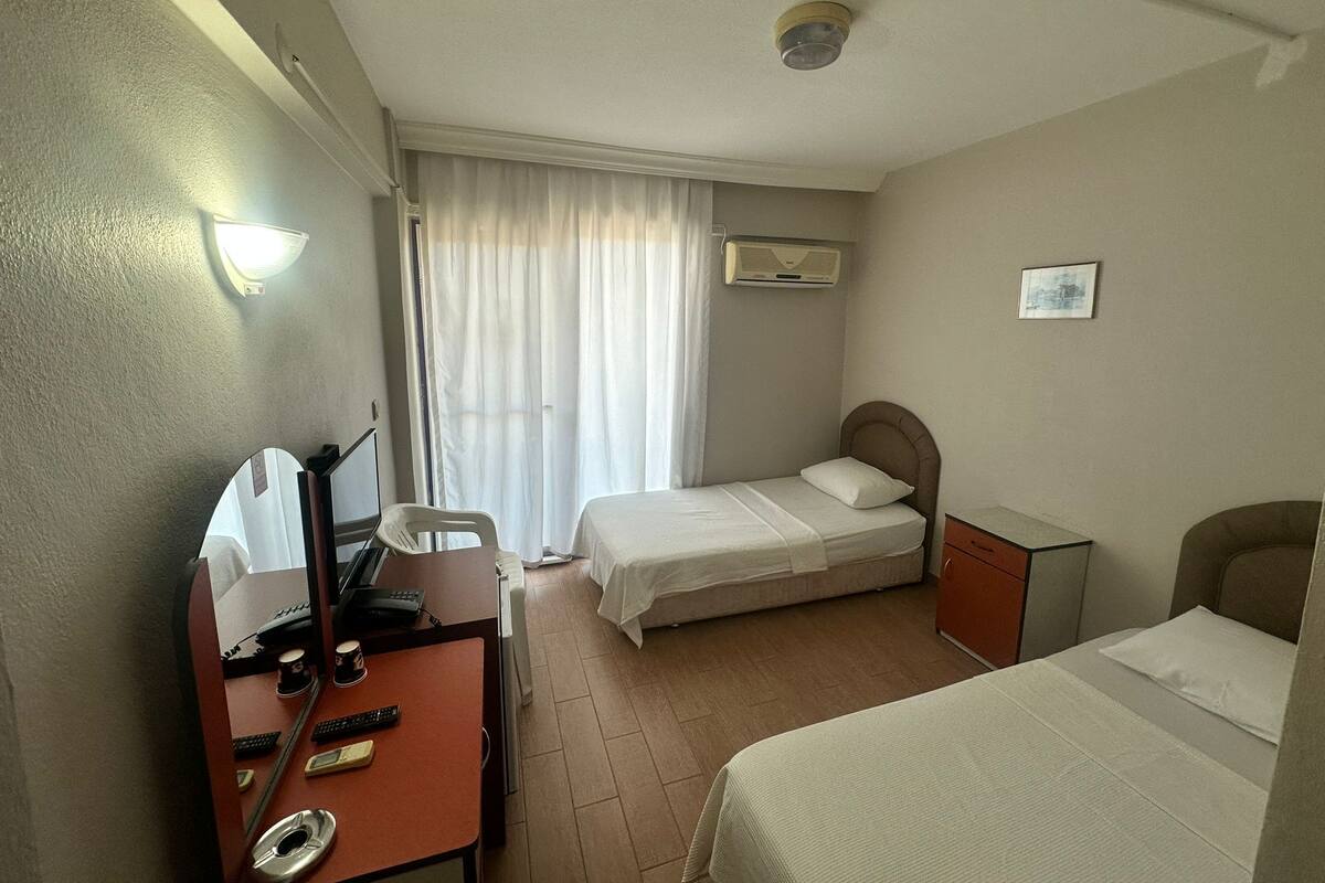 A hotel room with a desk, two beds, a chair, a window with curtains, an air conditioning unit, and a ceiling light.